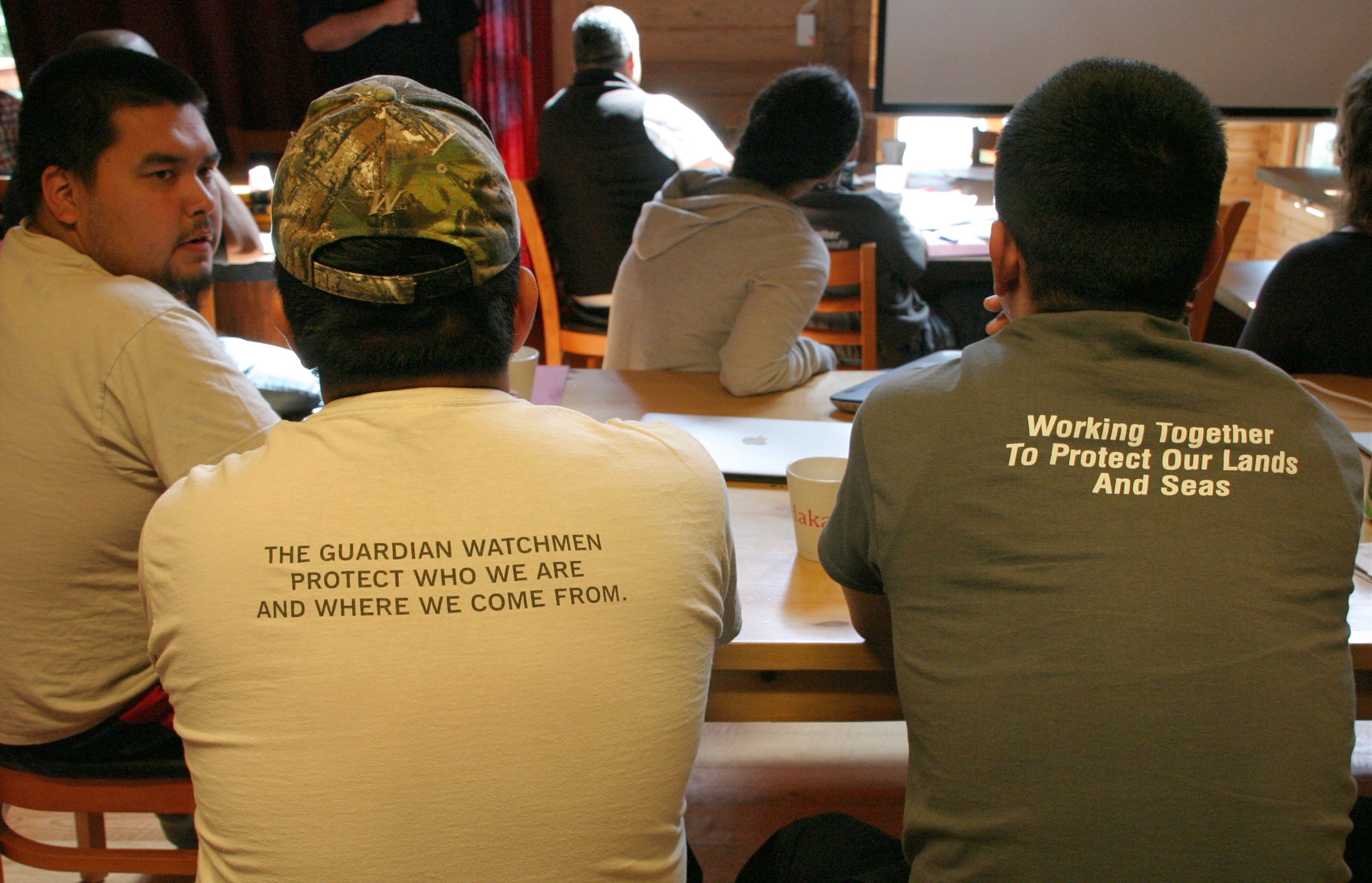 Two Coastal Guardian Watchmen showing the slogan on the back of their t-shirts, "The Guardian Watchmen protect who we are and where we come from." "Working together to protect our lands and seas."
